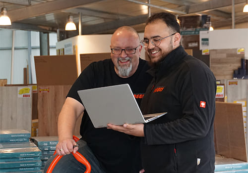 Kundenberatungsgespräch, ein Mitarbeiter hält dem Kunden einen Laptop hin, in den beide reinschauen, beide Personen lächeln, im Hintergrund ist die Filiale mit Produkten zu sehen